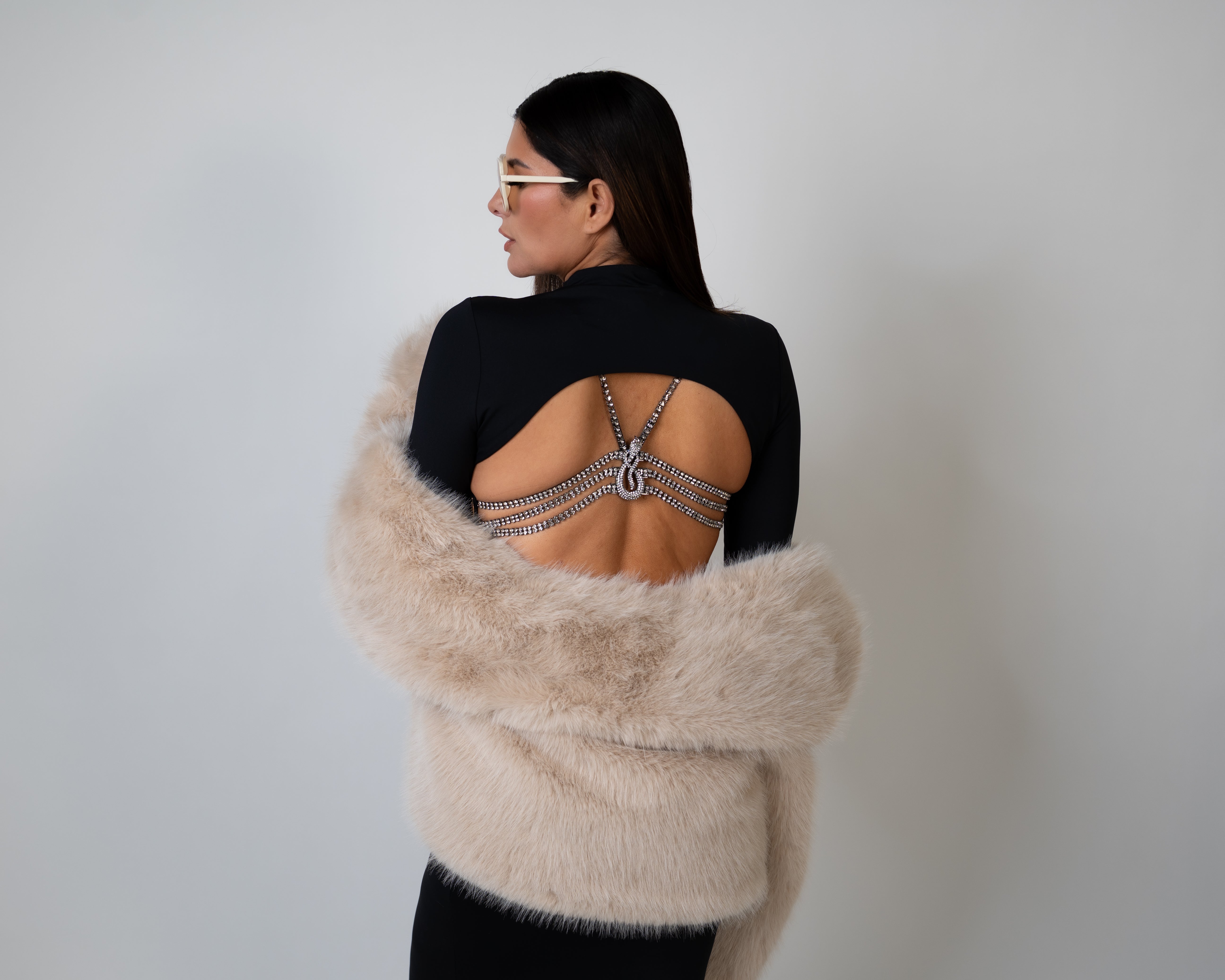 Woman wearing luxury lingerie bra on a backless dress wrapped in a beige fur coat against a plain background 