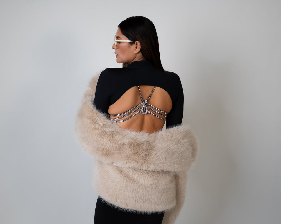 Woman wearing luxury lingerie bra on a backless dress wrapped in a beige fur coat against a plain background 